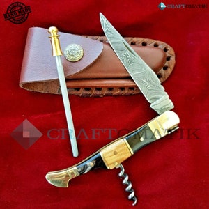 May include: A multi-tool pocket knife with a wood handle, a corkscrew, and a blade with a Damascus steel pattern. The knife is open and the blade is extended. The knife is in a brown leather sheath with a metal button. The knife is on a red velvet surface. The text "HAND MADE" is visible on a round stamp.