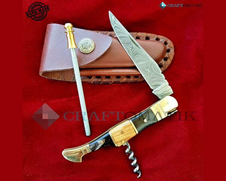 May include: A multi-tool pocket knife with a wood handle, a corkscrew, and a blade with a Damascus steel pattern. The knife is open and the blade is extended. The knife is in a brown leather sheath with a metal button. The knife is on a red velvet surface. The text "HAND MADE" is visible on a round stamp.