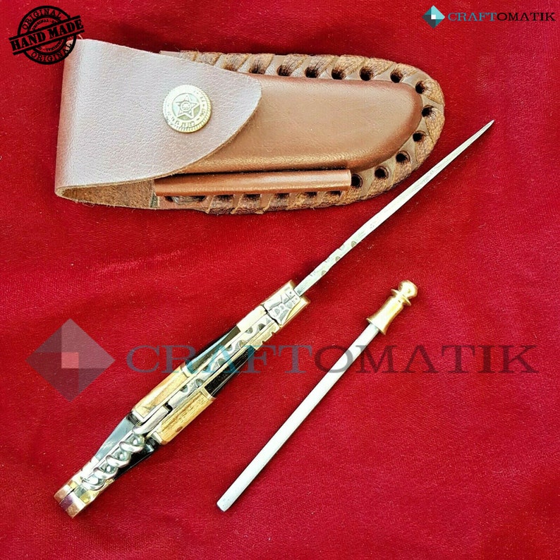 May include: A black and gold pocket knife with a decorative handle and a leather sheath. The knife has a small, round, gold-colored knob on the end of the blade. The sheath is brown leather with a decorative stitching pattern.