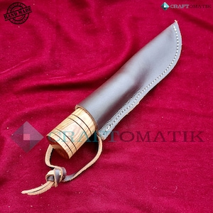 May include: A brown leather sheath with a wooden handle knife. The knife has a brown wooden handle with a light brown wood grain pattern. The sheath has a brown leather strap with a knot at the end. The knife is on a red velvet background.