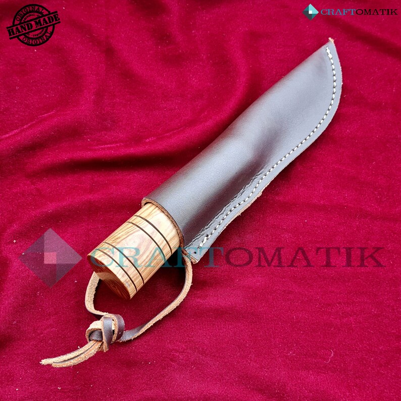 May include: A brown leather sheath with a wooden handle knife. The knife has a brown wooden handle with a light brown wood grain pattern. The sheath has a brown leather strap with a knot at the end. The knife is on a red velvet background.