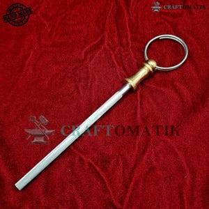 Könnte beinhalten: Ein silberner Schlüsselanhänger mit einem Messing-Schlüsselring und einer langen, dünnen, silbernen Klinge. Die Klinge ist mit einem Messing-Verbinder am Schlüsselring befestigt. Der Schlüsselanhänger befindet sich auf einem roten Samt-Hintergrund.