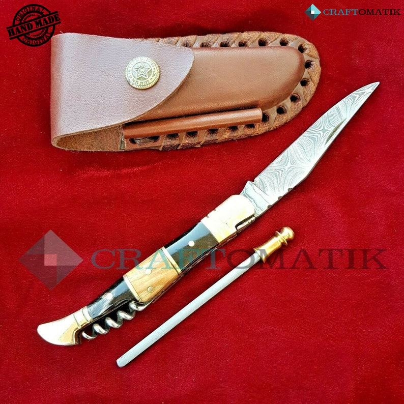 May include: A pocket knife with a wood and black handle, a corkscrew, and a metal rod. The knife has a patterned blade and comes with a brown leather sheath.