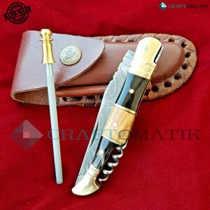 May include: A multi-tool pocket knife with a wood and black handle, a corkscrew, and a blade. The knife is open and has a damascus steel blade. The knife is in a brown leather sheath with a brass button.