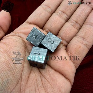 Könnte beinhalten: Drei quadratische Metalldürfel mit geätzten Mustern und nummerierten Flächen. Die Würfel werden in einer Hand gehalten, wobei die Zahlen 1 und 3 sichtbar sind. Die Würfel haben ein einzigartiges, strukturiertes Aussehen, und die Gesamtästhetik ist industriell.