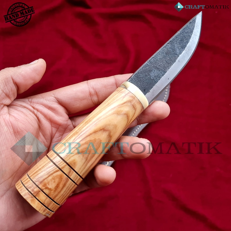 May include: A handmade knife with a wooden handle and a black blade. The handle has a light brown wood grain and is wrapped in a dark brown leather sheath. The blade is sharp and has a slight curve. The knife is a traditional design and is perfect for camping, hunting, or everyday use.