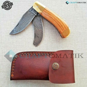 Puede incluir: Un cuchillo plegable de bolsillo con mango de madera y hoja de acero con patrón. El cuchillo está abierto y la hoja está extendida. El cuchillo viene con una funda de cuero marrón.