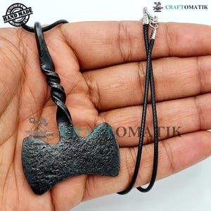 Puede incluir: Un colgante de hacha de metal negro con un mango retorcido, colgado de un collar de cordón negro. El colgante está forjado a mano y tiene un acabado rústico.
