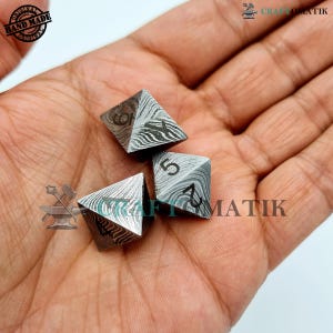 Könnte beinhalten: Drei Metallwürfel mit Pyramidenform und einer strukturierten Oberfläche. Die Würfel sind mit den Zahlen 4, 5 und 6 beschriftet. Die Würfel befinden sich in einer Hand.