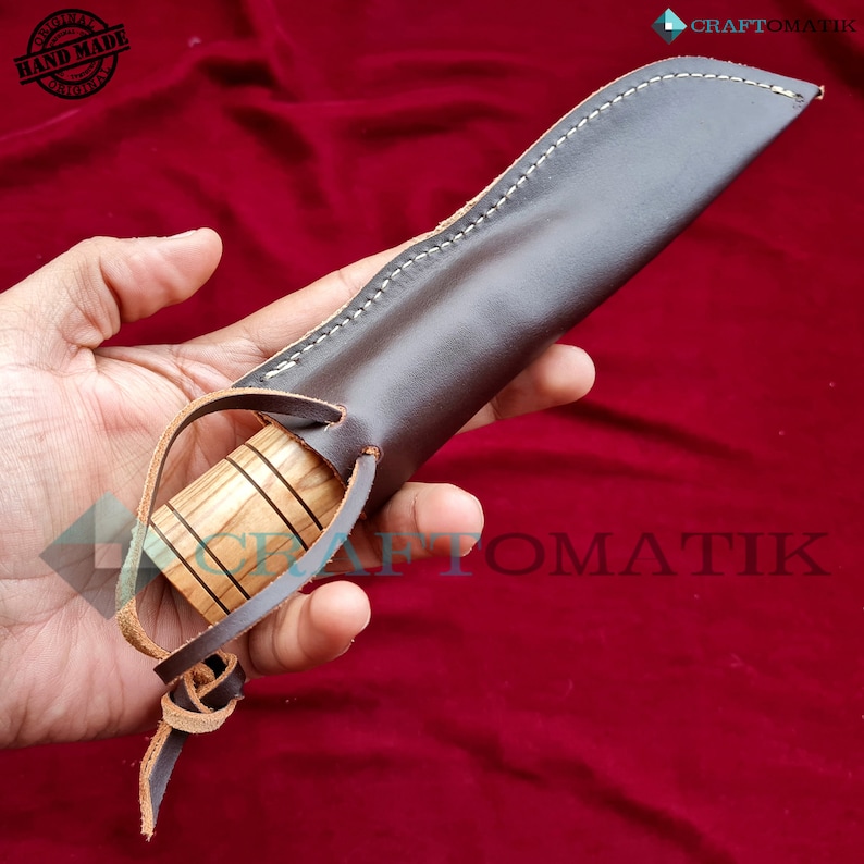 May include: A brown leather sheath with a stitched seam and a wooden handle. The sheath has a strap with a knot at the end. The sheath is held in a hand.