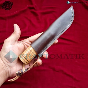 May include: A brown leather knife sheath with a wooden handle. The sheath has stitching around the edges and a leather strap. The handle is made of wood with a striped pattern. The knife is handmade and original.