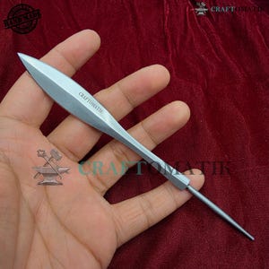 Pode incluir: Uma ferramenta pontiaguda prateada com o texto "CRAFTOMATIK" na lâmina. A ferramenta tem um cabo longo e fino e uma ponta triangular afiada. A ferramenta é segurada em uma mão contra um fundo vermelho escuro.