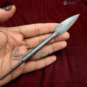 May include: A silver spearhead with a patterned blade, held in a hand against a burgundy background. The spearhead has a pointed tip and a long, slender shaft. The blade has a unique, textured design.