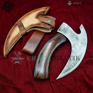 Könnte beinhalten: Eine geschwungene Axt aus Metall mit einem Griff aus dunklem Holz und geätzten Mustern auf der Klinge, begleitet von einer hellbraunen Lederscheide mit einem Messingdruckknopf. Die Klinge zeigt komplexe Gravuren und Runensymbole.