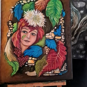 Puede incluir: Pintura original sobre lienzo que representa el rostro de una mujer rodeado de hojas coloridas y una flor blanca. La obra presenta un arco de piedra y un fondo marrón, expuesta sobre un caballete de madera.