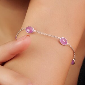 May include: A silver bracelet with oval, lilac-coloured gemstones. The bracelet is displayed on a wrist, with the gemstones spaced along the chain. The design is simple and elegant, suitable for everyday wear. The gemstones are approximately 0.5 cm in diameter.