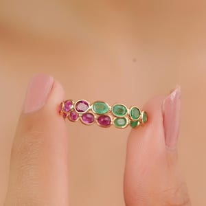 Può includere: Un anello d'oro con pietre preziose ovali rosa e verdi alternate. L'anello è tenuto tra due dita, mettendo in risalto i colori vivaci e il design intricato. Le pietre sono incastonate vicine, creando una fascia continua di colore.