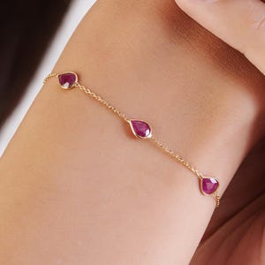 Könnte beinhalten: Ein zartes Armband aus einer goldfarbenen Kette mit drei gleichmäßig verteilten, birnenförmigen, tiefroten Rubin-Edelsteinen. Das Armband wird durch eine dünne, polierte Goldgliederkette gesichert, die sich um das Handgelenk legt und die leuchtend roten Steine präsentiert.