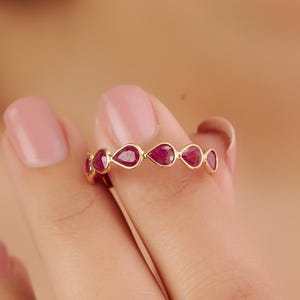 Op de afbeelding: Een gouden ring met een reeks druppelvormige, dieprode edelstenen. De ring wordt omhoog gehouden, waardoor de kleur van de edelstenen en het delicate ontwerp van de ring worden benadrukt. De ring is een sieraad.
