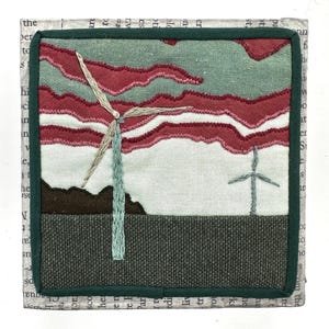 Pode incluir: Peça de arte têxtil bordada com uma paisagem com duas turbinas eólicas. O céu é retratado com nuvens costuradas vermelhas e verdes. A obra de arte é emoldurada com uma borda verde e um fundo de texto impresso.