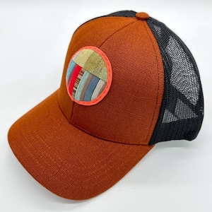 May include: A rust-colored baseball cap with a black mesh back. The front panel features a circular patch with a multi-colored vertical striped pattern in shades of green, red, blue, and tan, outlined by a bright neon orange stitched border.