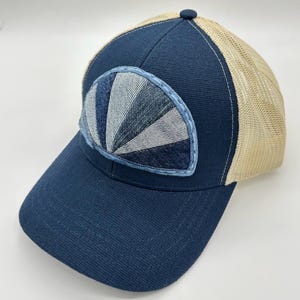 May include: A navy blue baseball cap with a beige mesh back and a structured front panel. The front features a semi-circular patch with a sunburst pattern made of alternating light and dark blue denim fabric segments with stitched edges.