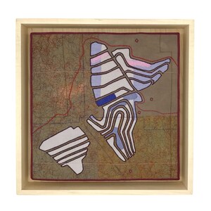 Op de afbeelding: Een ingelijst textielkunstwerk dat een kaart toont met een gestileerd ontwerp van een landschap in tinten blauw, roze en wit. De kaart is omgeven door een bruin frame.