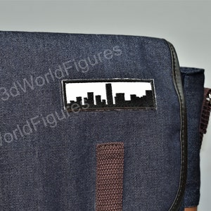 May include: A dark blue denim messenger bag with a brown strap and a white patch featuring a black silhouette of a city skyline.