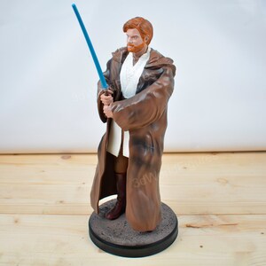 Pu&ograve; includere: Statuetta di un personaggio di un franchise di fantascienza. La figura ha i capelli rossi e una veste marrone, che tiene una spada laser blu. La figura &egrave; su una base rotonda grigia.
