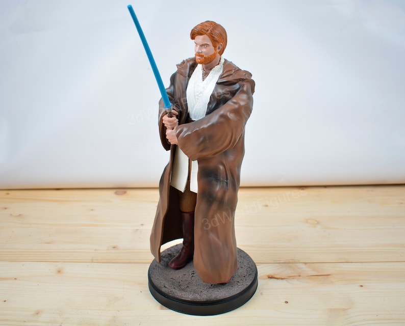 Pu&ograve; includere: Statuetta di un personaggio di un franchise di fantascienza. La figura ha i capelli rossi e una veste marrone, che tiene una spada laser blu. La figura &egrave; su una base rotonda grigia.