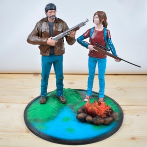 Pu&ograve; includere: Un set di figurine dettagliate raffiguranti due personaggi, uno con un fucile a canna liscia e l'altro con un fucile. Si trovano su una base circolare con un paesaggio dipinto, tra cui un fal&ograve; e acqua. Le figure sono vestite in modo casual.