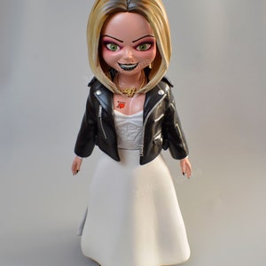 Pu&ograve; includere: Statuetta della bambola Chucky con giacca di pelle nera e abito da sposa bianco. La bambola ha i capelli biondi, occhi verdi e un sorriso sinistro. Sono visibili anche una collana d'oro e un tatuaggio a forma di cuore.