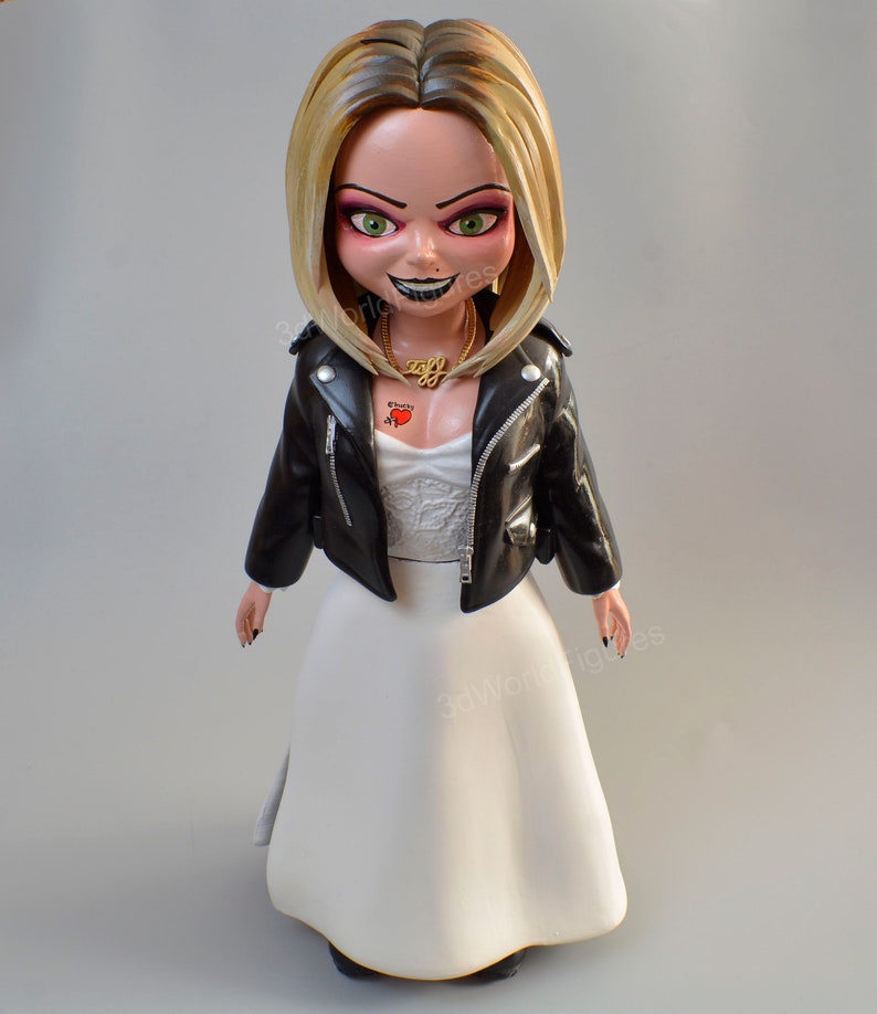 Pu&ograve; includere: Statuetta della bambola Chucky con giacca di pelle nera e abito da sposa bianco. La bambola ha i capelli biondi, occhi verdi e un sorriso sinistro. Sono visibili anche una collana d'oro e un tatuaggio a forma di cuore.