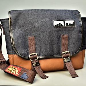 May include: A blue denim messenger bag with brown leather accents and a brown strap. The bag has a patch with a cityscape design and a red and yellow patch that says "Cool Story".