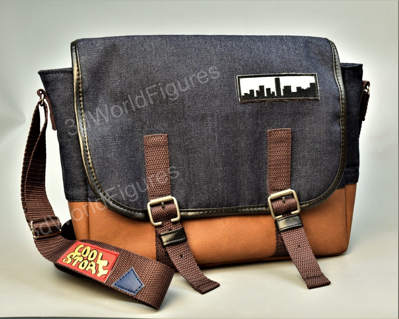 May include: A blue denim messenger bag with brown leather accents and a brown strap. The bag has a patch with a cityscape design and a red and yellow patch that says "Cool Story".