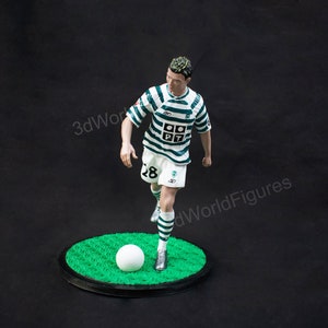 Pu&ograve; includere: Statuetta di un calciatore con maglia a righe verdi e bianche e pantaloncini bianchi, che calcia un pallone bianco. La figura &egrave; su una base circolare verde con bordo nero, su sfondo scuro.