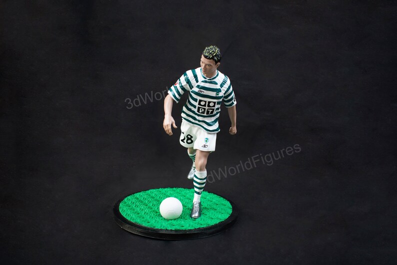 Pu&ograve; includere: Statuetta di un calciatore con maglia a righe verdi e bianche e pantaloncini bianchi, che calcia un pallone bianco. La figura &egrave; su una base circolare verde con bordo nero, su sfondo scuro.