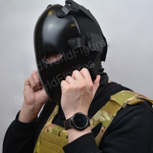 May include: A person wearing a black tactical mask with a black balaclava and a camouflage vest. The person is adjusting the mask with their hands. The person is wearing a black smartwatch on their left wrist.