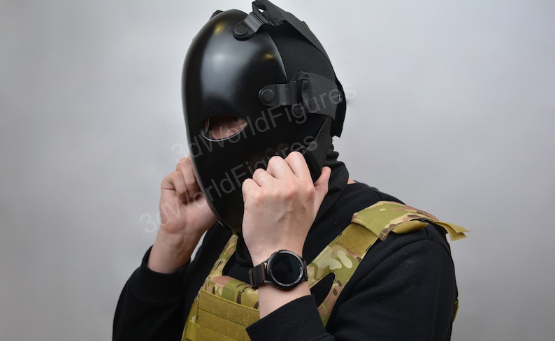 May include: A person wearing a black tactical mask with a black balaclava and a camouflage vest. The person is adjusting the mask with their hands. The person is wearing a black smartwatch on their left wrist.