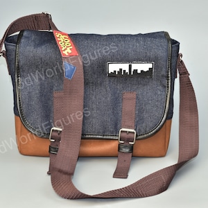 May include: A blue denim messenger bag with brown leather accents and a brown adjustable strap. The bag has a white city skyline patch and a yellow and red patch that says "Cool Spot".