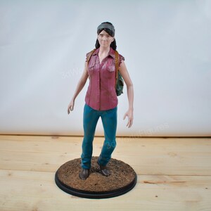 Pu&ograve; includere: Statuetta dettagliata di una persona con una camicia bordeaux abbottonata, jeans blu e una fascia. La figura ha uno zaino e si trova su una base circolare. La base &egrave; marrone con un bordo nero.