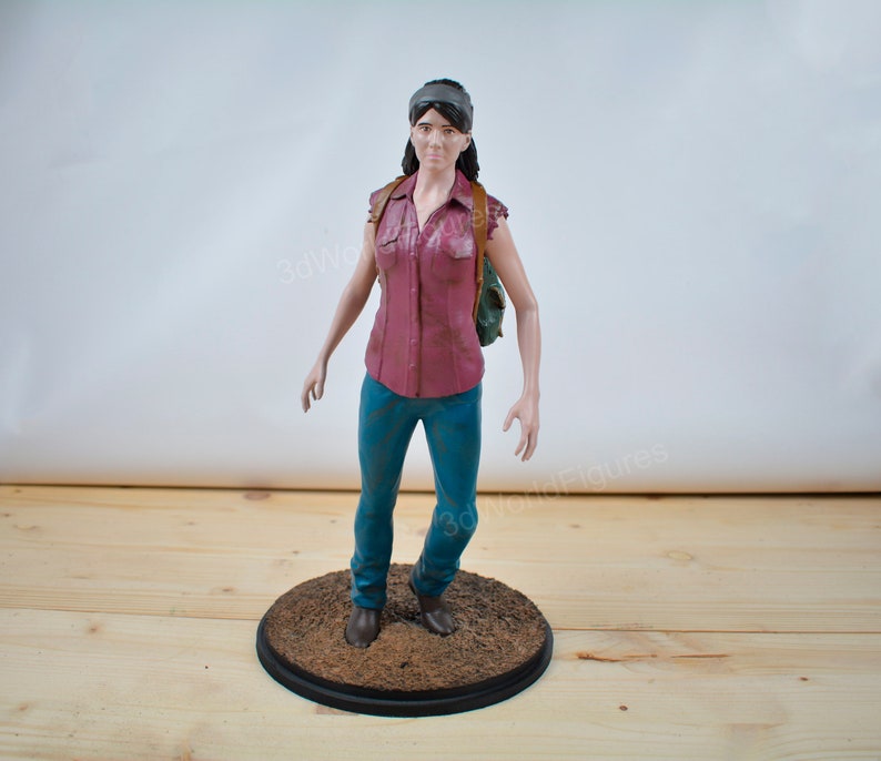 Pu&ograve; includere: Statuetta dettagliata di una persona con una camicia bordeaux abbottonata, jeans blu e una fascia. La figura ha uno zaino e si trova su una base circolare. La base &egrave; marrone con un bordo nero.