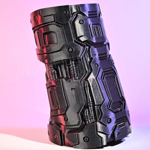 Puede incluir: Un guantelete negro de aspecto futurista con un diseño mecánico complejo. El guantelete presenta varios paneles, remaches y lo que parecen ser muelles, lo que le da una estética robusta e industrial. El objeto está inclinado sobre un fondo degradado rosa y morado.
