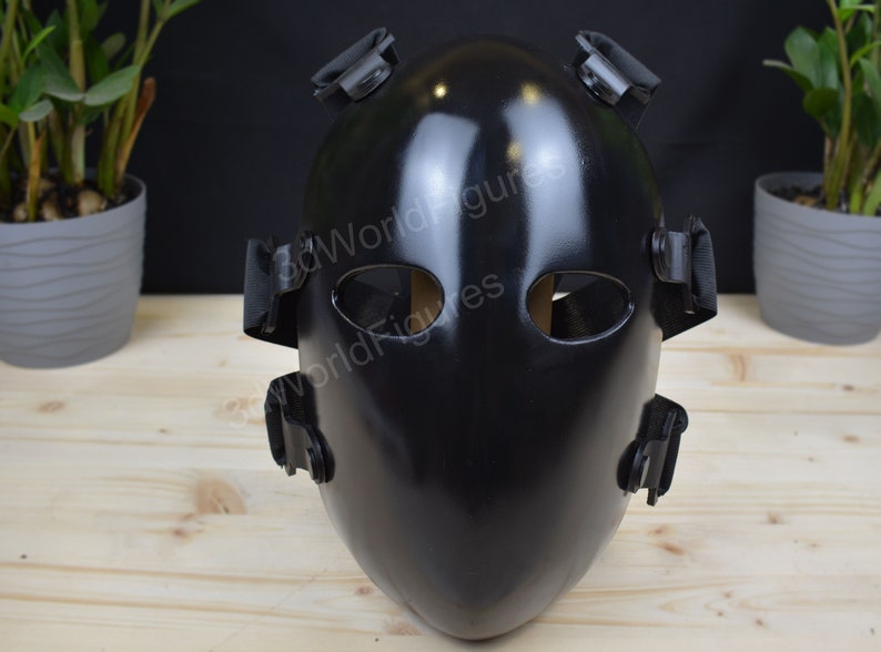 May include: A black protective face mask with adjustable straps. The mask has two eye holes and a smooth, glossy finish.