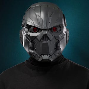 May include: A matte dark gray full-face helmet featuring a complex mechanical design with angular panels, recessed vents, and two glowing red circular eye lenses. The helmet includes integrated side circular components and small tubing details.