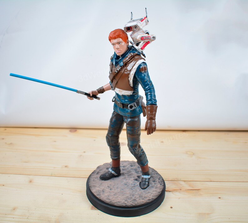 Pu&ograve; includere: Statuetta dettagliata di un personaggio di un franchise di fantascienza, con una spada laser blu. La figura indossa un abito blu e marrone, con un piccolo droide sulla schiena. La statuetta &egrave; su una base circolare.