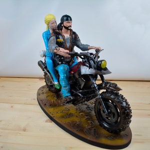 Pu&ograve; includere: Statuetta dettagliata di una coppia in moto. L'uomo indossa berretto e giacca neri, mentre la donna ha i capelli biondi e abiti blu. La moto &egrave; scura con sella rossa, su base marrone.