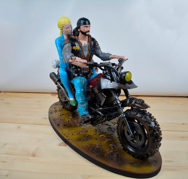 Pu&ograve; includere: Statuetta dettagliata di una coppia in moto. L'uomo indossa berretto e giacca neri, mentre la donna ha i capelli biondi e abiti blu. La moto &egrave; scura con sella rossa, su base marrone.