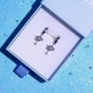 Puede incluir: Un par de pendientes de aro plateados con un diseño celestial, presentados en una caja azul claro. Cada pendiente presenta una parte superior en forma de estrella con cristales transparentes y un dije de estrella colgante. La caja tiene un interior blanco y una cinta azul.