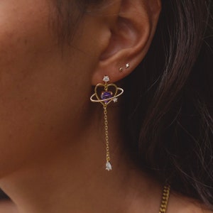 May include: Gold heart-shaped earrings with a purple gemstone and a dangling chain. The earrings feature a small circle around the heart, with three small clear gemstones.